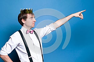 Man with dark hair in pink shirt, bow tie, suspender and gold shiny homemade crown pointing with finger to away