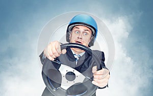 Man in a dark suit and a motorcycle helmet holds a car steering wheel in his hands while making a turn, with smoke