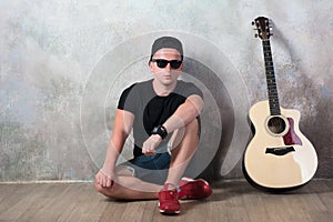 Man in denim shorts sitting next to a guitar on the wall background in style grunge, music, musician, hobby, lifestyle, hobby