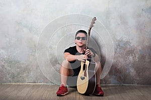 Man in denim shorts sitting next to a guitar on the wall background in style grunge, music, musician, hobby, lifestyle, hobby