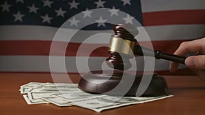 Man at desk with wooden gavel, dollar bills and US flag in background. A judge in a courtroom, striking with a wooden