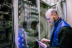 A man with a digital tablet working in a server room. The engineer stands next to a multitude of internet wires. The technician