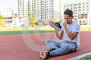 A man disgusted by the smell of his running shoe. A sport man sitting on the  track race at stadium