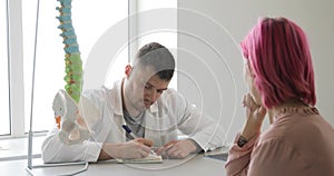 Man doctor and patient woman with pink hairs are discussing diagnosis using spine model.