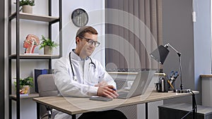 Man doctor talking to online patient on laptop screen sitting at clinic office desk giving online consultation