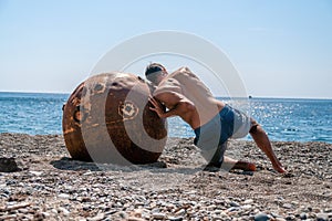 Man doing exercise outdoors near to old rusty floating marine mine on the beach with rocky shore and sea background