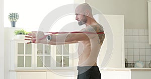 Man doing fitness exercise for shoulders using elastic band at home, side view.