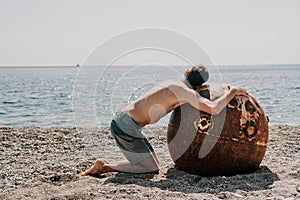 Man doing yoga and meditation outdoors near to old rusty floating marine mine on the beach with rocky shore and sea