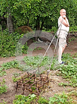 A man drags an old harrow by a rope in the yard with an effort. Rural life