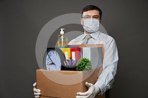 A man dressed as a businessman holds a box with office stuff, documents, hand sanitizer, posing in studio on gray background,
