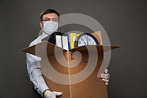 A man dressed as a businessman holds a box with office stuff, documents, posing in studio on gray background, medical face mask