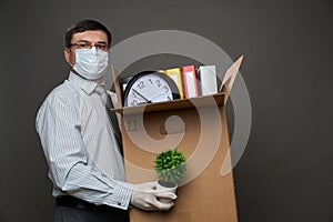 A man dressed as a businessman holds a box with office stuff, documents, posing in studio on gray background, medical face mask