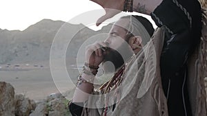 Man dressed as a shaman, standing on a rock in the middle of the desert sands and playing the harmonica