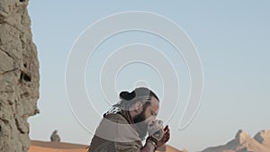 Man dressed as a shaman, standing on a rock in the middle of the desert sands and playing the harmonica