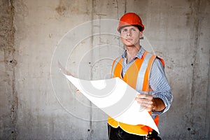 Man dressed in orange work vest and helmet explores construction documentation on a concrete wall background