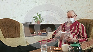 A man in a dressing gown and a medical mask in quarantine self-isolation writes in a notebook on a sofa.