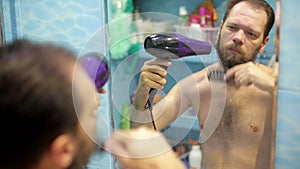 a man dries his beard with a hair dryer after washing in the bathroom in front of a mirror