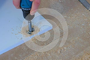 Man drill round hole for door hinges in chipboard usinf electric drill, closeup.