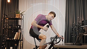 Man is drinking water while cycling on indoor smart bike trainer. Indoor cycling. Cyclist is training on exercise bicycle at home.