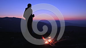 A man drinks a hot drink at night by a campfire in a tourist camp on a mountain pass. A tourist admires the beautiful views of the