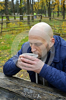 A man drinks tea from a mug while sitting at a table in an autumn park