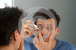 Man is dripping drops in his eyes in front of the mirror at home. photo