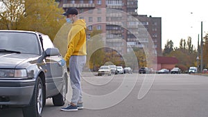 A man, a driver in disposable gloves and a medical mask, opens the driver's seat door, gets behind the wheel of a car