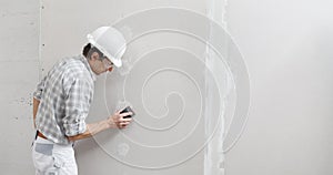 Man drywall worker or plasterer sanding and smoothing a plasterboard walls with stucco using a sandpaper holder. Wearing white