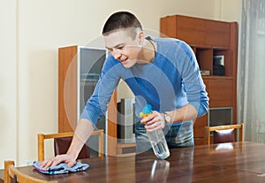 Man dusting wooden table with rag and cleanser at home