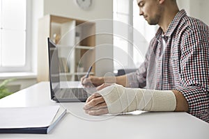 Man with elastic bandage on his hand sitting at table, working on laptop and writing in notebook