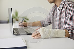 Man with elastic bandage wrapped around his wrist sitting at desk and working on laptop