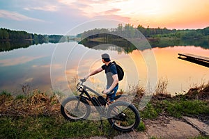 Man with an electric fatbike on the background of a lake and sunset. Picturesque place in the village. Concept of a healthy