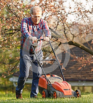 Man with electric lawnmower, lawn mowing. Gardener trimming a garden. Sunny day, suburb, village.