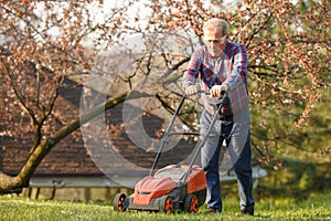 Man with electric lawnmower, lawn mowing. Gardener trimming a garden. Sunny day, suburb, village. Adult man pruning and