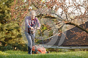 Man with electric lawnmower, lawn mowing. Gardener trimming a garden. Sunny day, suburb, village. Adult man pruning and