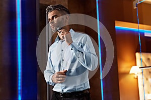 Man With Electric Razor Shaving Face In Bathroom. Men Grooming