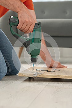 Man with electric screwdriver assembling furniture on floor indoors, closeup