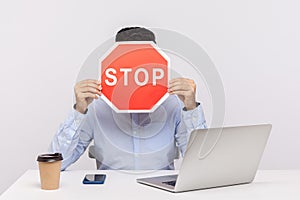Man employee sitting in office workplace with laptop, hiding face behind stop symbol, warning with red traffic sign