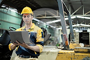 Man engineer wear yellow hard hat and wearing safety uniform standing at forklift using laptop working at heavy industrial factory