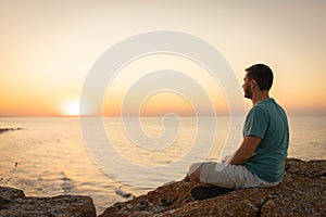 A man enjoys the view of the sunset on the sea, sitting on a rock.