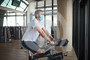 Man exercising in the gym on stationary bike indoor cycling wearing face mask to prevent coronavirus