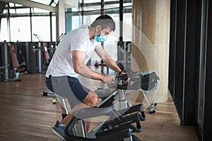 Man exercising in the gym on stationary bike indoor cycling wearing face mask to prevent coronavirus