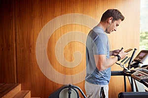 Man Exercising On Treadmill At Home Wearing Wireless Earphones Checking Mobile Phone