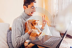 Man facetiming from home holding pet using computer. Ginger cat looking at laptop screen during video chat.