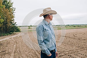 Man farmer agronomist in jeans and shirt stands back in field after haymaking  with tablet looking into the distance. Rural