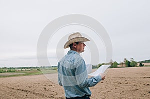 Man farmer agronomist in jeans and shirt stands back in field after haymaking  with tablet looking into the distance. Rural