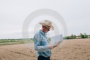 Man farmer agronomist in jeans and shirt stands back in field after haymaking  with tablet looking into the distance. Rural