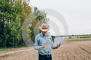 Man farmer agronomist in jeans and shirt stands back in field after haymaking  with tablet looking into the distance. Rural