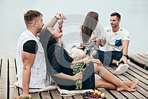 Man feeding his girlfriend grapes sitting on wooden pier on picnic