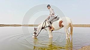 man is feeding water to thirsty horse from pond or Horse drinking water in river while men seated on it